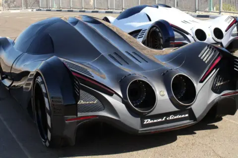 Dwa futurystyczne samochody Devel Sixteen, jeden czarny, drugi biały, stoją na asfalcie.