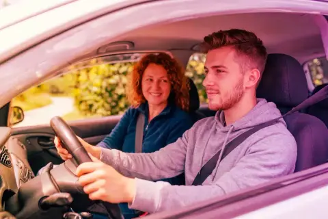 Giovane uomo guida, madre sorride. Ricorda che l'assicurazione auto deve essere intestata al proprietario per la massima tranquillità.