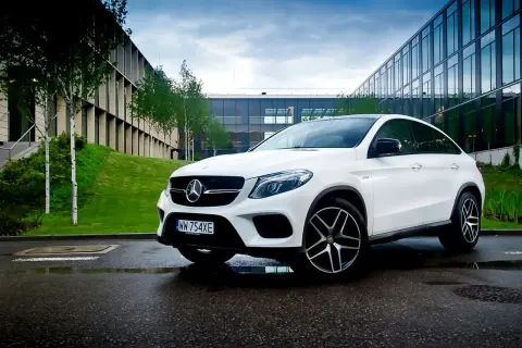 Biały Mercedes GLE 450 zaparkowany na mokrym asfalcie przed nowoczesnym budynkiem.