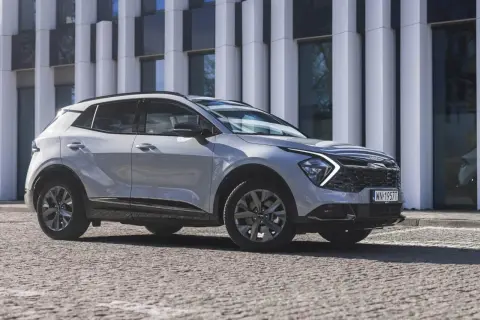 Nowy Kia Sportage Anniversary w jasnym kolorze, zaparkowany przed nowoczesnym budynkiem. Stylowy SUV na kamienistej nawierzchni.