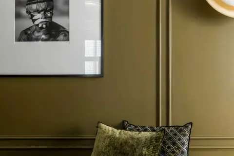 Un pan de mur vert olive, orné d'un portrait encadré d'un jeune homme au visage peint, et de coussins décoratifs.