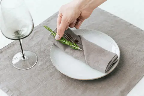 Une main dépose une branche de romarin sur un pliage de serviette mariage boheme, sur une assiette blanche.