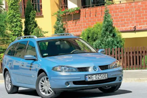 Błękitne kombi Renault Laguna 2 z relingami dachowymi stoi przed domem z żółtą elewacją i drewnianym płotem.