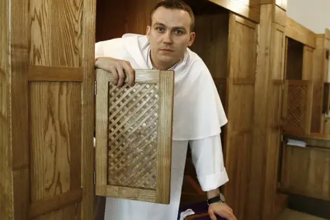 Ksiądz w białej albie otwiera kratkę konfesjonału, czekając, kiedy jest spowiedź w kościele.