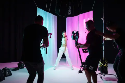 Filming a music video, learning how to color lighting with a dancer posed between blue and pink backdrops, while camera operators film.