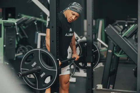 Mann mit Bandana macht schwere myo reps mit Langhantel im Fitnessstudio.