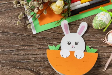 Uroczy króliczek z papieru kolorowego wychyla się z marchewki. Obok pisanki z papieru kolorowego, bazie i flamastry.