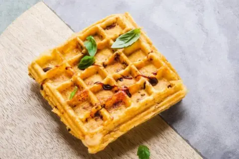 Goldbraune belgische Waffeln mit dunklem Sirup und frischem Basilikum, serviert auf einem Holzbrett.