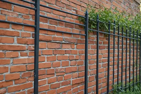 Czerwone, ceglane ogrodzenie z czarnymi, metalowymi prętami i zielonymi pnączami.