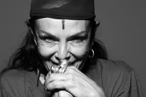 Michele Lamy, con un turbante nero e un segno sulla fronte, sorride enigmaticamente, le mani adornate da anelli vistosi.