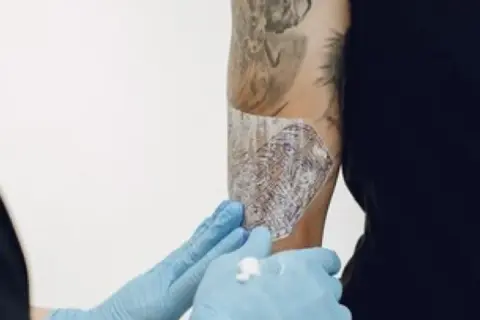 Manos enguantadas aplican Bepanthol sobre un tatuaje para ayudar a su curación.