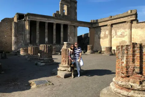 Pareja posa frente a ruinas antiguas, preguntándose donde está Pompeya. El sol ilumina columnas rotas y edificios históricos.