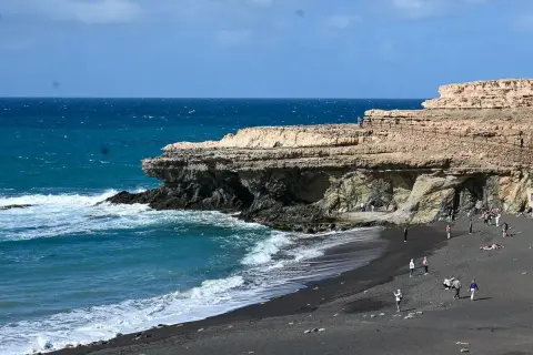 Fuerteventura Urlaub Tipps: Schwarzer Strand mit Felsen und türkisblauem Meer. Menschen spazieren und entspannen.