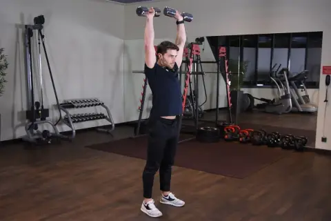 Wyciskanie hantli nad głowę angażuje mięśnie naramienne i triceps. Mężczyzna wykonuje ćwiczenie w siłowni.