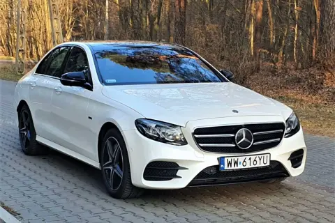 Biały Mercedes E300e z czarnym dachem i lusterkami, zaparkowany na brukowanej ścieżce w lesie.
