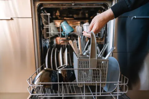 Ręka wkłada koszyk ze sztućcami do zmywarki. To oznacza, że naczynia są "dishwasher safe", czyli można je myć w zmywarce.