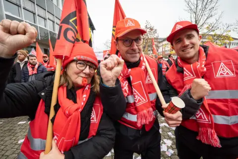 Drei Personen in roten Westen und Schals mit Gewerkschaftssymbolen, die Fahnen schwenken und die Faust ballen.