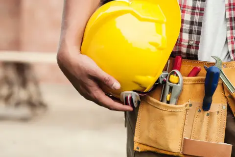 Un trabajador sostiene un casco amarillo. Su cinturón de herramientas tiene destornilladores y una llave inglesa. Esto es lo que es el ITE: seguridad y preparación.