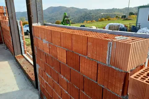 Muro in costruzione con blocchi di laterizio, un materiale che resiste al fuoco, con vista su colline verdi e un escavatore.