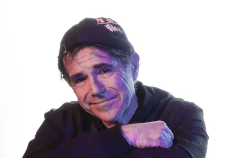 Andrea Satta, con un sorriso enigmatico, indossa un cappellino scuro e una felpa. La luce colorata crea un'atmosfera suggestiva.