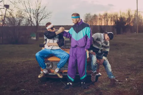 Trzech mężczyzn w dresach i swetrach w stylu lat 90. siedzi na pomarańczowym samochodzie.