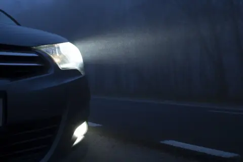 El coche avanza en la niebla con sus luces antiniebla encendidas, iluminando el camino oscuro y los árboles a un lado.
