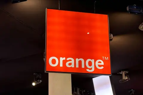 Jasny, pomarańczowy znak z napisem "orange" wisi pod sufitem. To logo Orange, które często pojawia się na ekranach telewizorów.