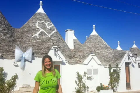 Una donna sorride davanti ai trulli di Alberobello, patrimonio UNESCO, con un grande fiocco azzurro appeso a un cono.