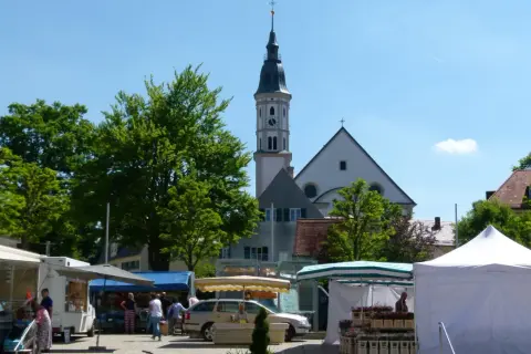 Bunter Markt in Mering mit Kirche im Hintergrund. Stände mit Blumen und Lebensmitteln, Menschen bummeln.