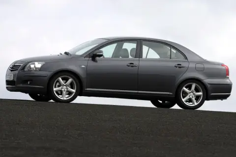 Szary sedan Toyota Avensis 1.8 benzyna stoi na ciemnej nawierzchni, z niebieskim niebem w tle.