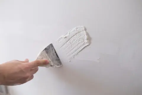 Dłoń z kielnią nakłada gips szpachlowy na białą ścianę.