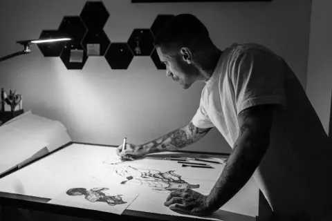 Víctor del Fueyo, artista con tatuajes, dibuja con concentración bajo una lámpara.