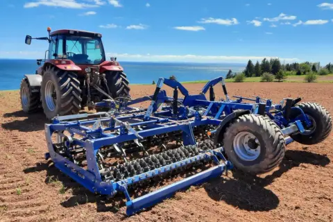 Czerwony traktor z niebieskim agregatem uprawowym na polu, gotowy do siewu. W tle błękitne niebo i morze.