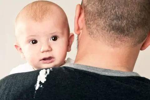 En bebis med stora ögon tittar fram över pappans axel, som har fått en kladdig fläck.