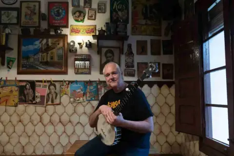Un hombre sonriente con un banjo en salsipuedes una tasca encantadora, rodeado de carteles vintage y objetos curiosos.