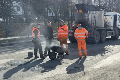 Ekipa drogowców w pomarańczowych strojach pracuje przy drodze, obok wózka i specjalistycznej ciężarówki.