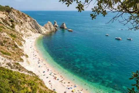 Spiaggia affollata ai piedi del Monte Conero, con acque cristalline e barche all'orizzonte.