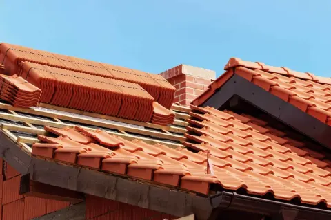 Układanie ceramicznych dachówek na dachu w budowie, widoczny komin i niebo.