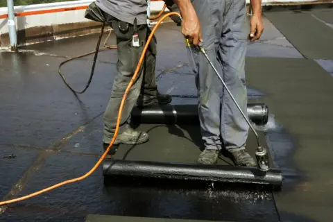 Zwei Dachdecker arbeiten auf einem Flachdach. Einer hält einen Brenner, um die Dachpappe zu schweißen.