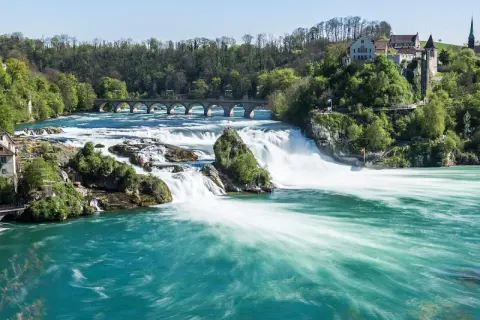 Breite, türkise Wasserströme stürzen über Felsen, umgeben von grünen Hügeln mit Häusern und einer Brücke. Die Rhine Waterfalls sind ein beeindruckendes Naturschauspiel.