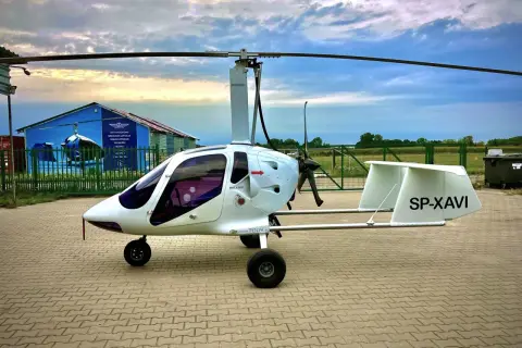 Biały wiatrakowiec SP-XAVI gotowy do lotu. Na tle chmur i niebieskiego hangaru z napisem "Loty widokowe".