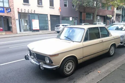 Klasyczny beżowy BMW E10 z lat 70. stoi zaparkowany przy ulicy, obok nowszego srebrnego samochodu.