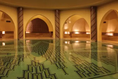 Wnętrze hammamu z basenem o geometrycznym wzorze na dnie. Wokół łuki, kolumny zdobione mozaiką i kamienne ławy.