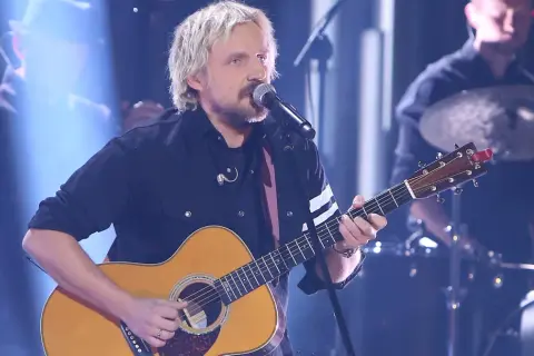 Artysta z blond włosami gra na gitarze akustycznej, śpiewając do mikrofonu. Juz teraz wiem ze dni tekst.
