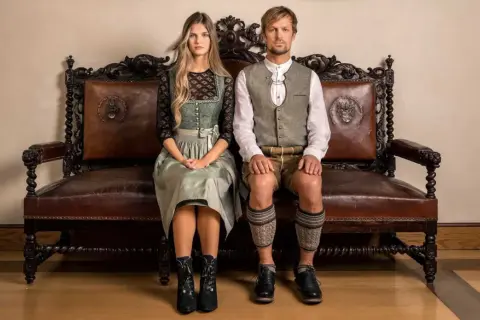 Ein junges Paar sitzt in traditioneller Tracht auf einer antiken Ledercouch. Sie sind bereit, in Tracht zu heiraten und zeigen stolz ihre festliche Kleidung.