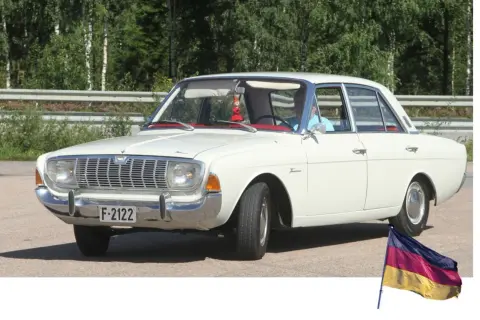 Vit Ford Taunus, en klassisk modell av svenska bilar, parkerad utomhus med en tysk flagga i förgrunden.