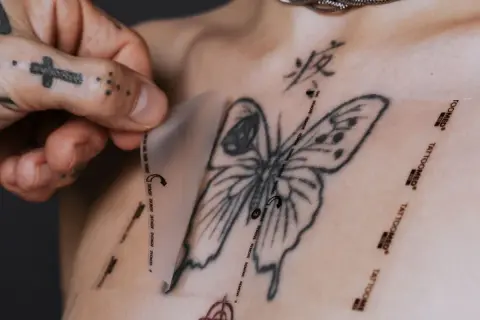 Ein Mann mit tätowierter Hand zieht eine Schutzfolie von einem frisch gestochenen Schmetterling-Tattoo auf der Brust ab. Pflege danach ist wichtig.