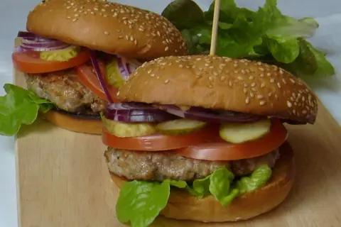 Dwa soczyste burgery z bułkami do hamburgerów pysznie czy przepysznie, z pomidorami, ogórkami, cebulą i sałatą.