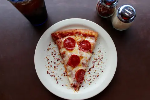 Kawałek pizzy pepperoni na talerzu, obok słoiczki z przyprawami i napój. Zastanawiasz się, ile kosztuje pizza we Włoszech?