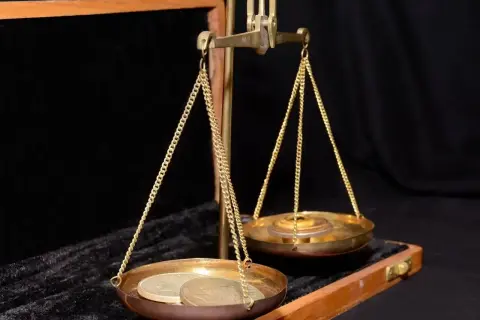 Waga szalkowa z monetami i ciężarkami. Na jednej szalce leżą monety, na drugiej ciężarki, symbolizujące uncję złota.